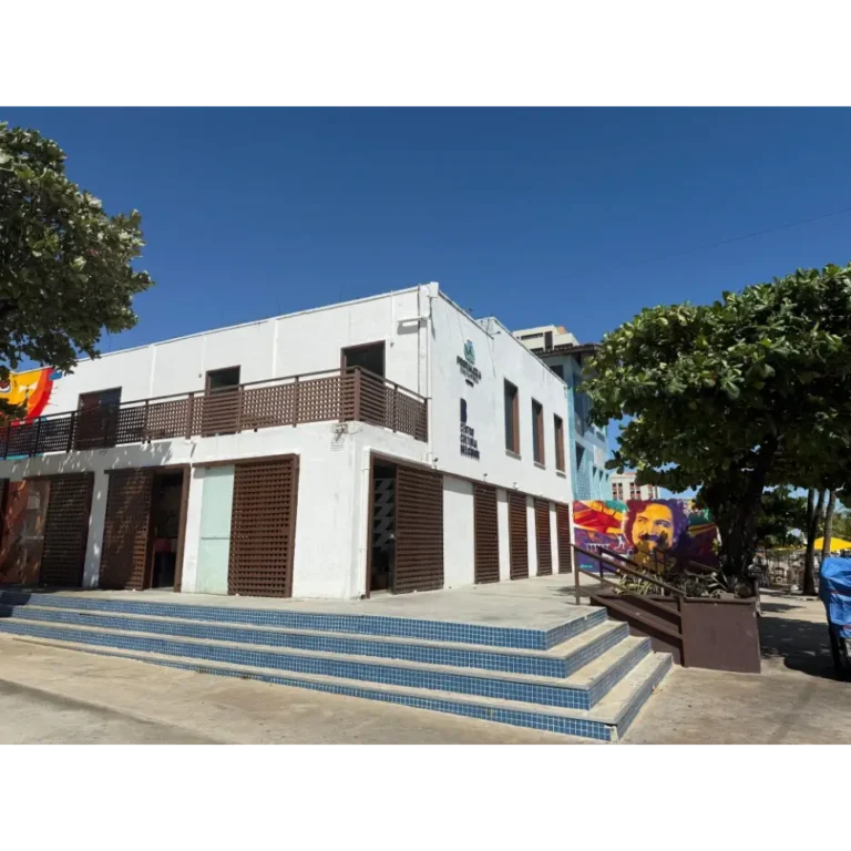 Vista frontal do Centro Cultural Belchior em Fortaleza, um prédio branco de dois andares com detalhes em madeira e escadaria azul, localizado próximo à Praia de Iracema sob um céu azul