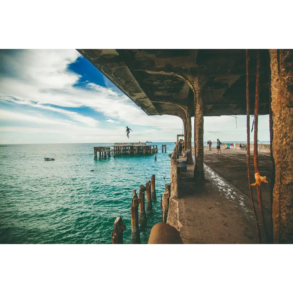 Fotografia artística tirada debaixo da estrutura da Ponte Metálica, mostrando uma pessoa saltando no mar azul, com pilares de madeira e a silhueta da ponte emoldurando a cena.