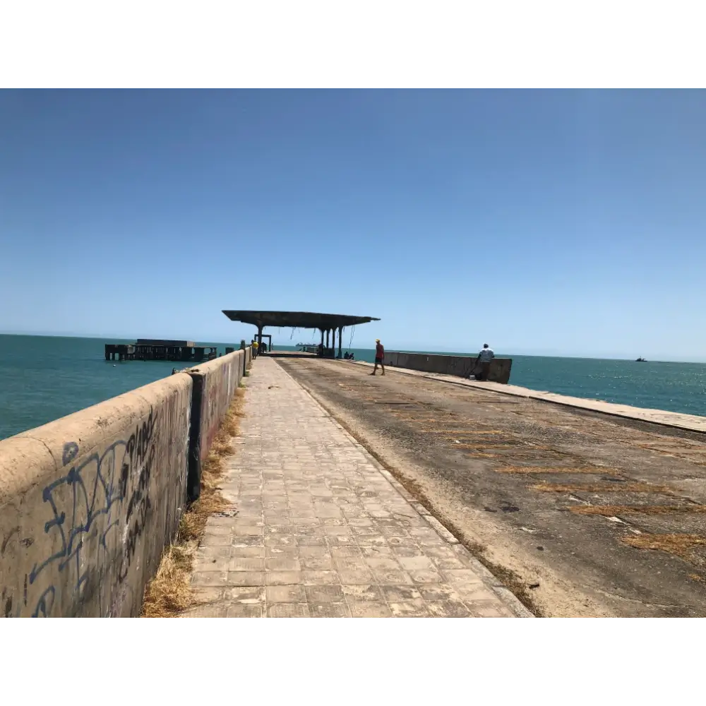 Perspectiva de cima da Ponte Metálica no Poço da Draga, mostrando o piso de concreto com marcas de ferrugem, a mureta lateral com grafites e o mar azul ao fundo.