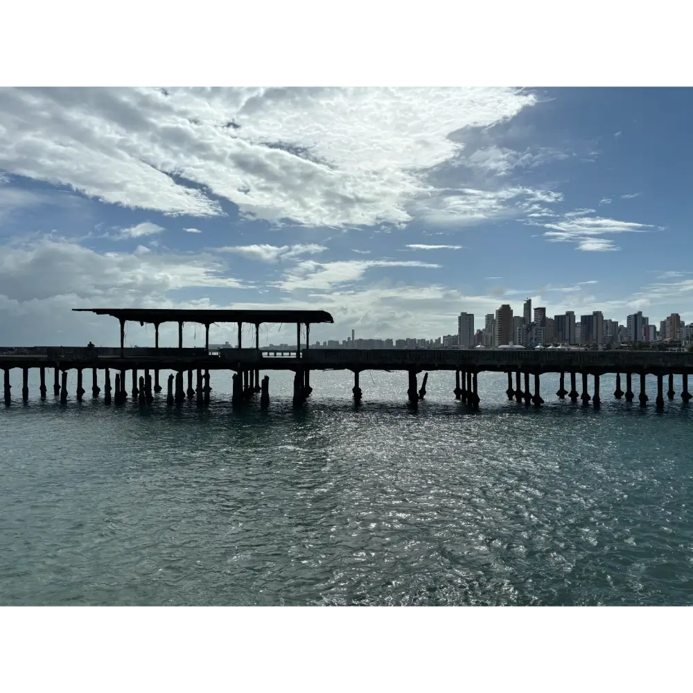 Vista lateral da Ponte Metálica no Poço da Draga em Fortaleza, com o mar em primeiro plano e os prédios da orla da cidade ao fundo sob um céu com nuvens.