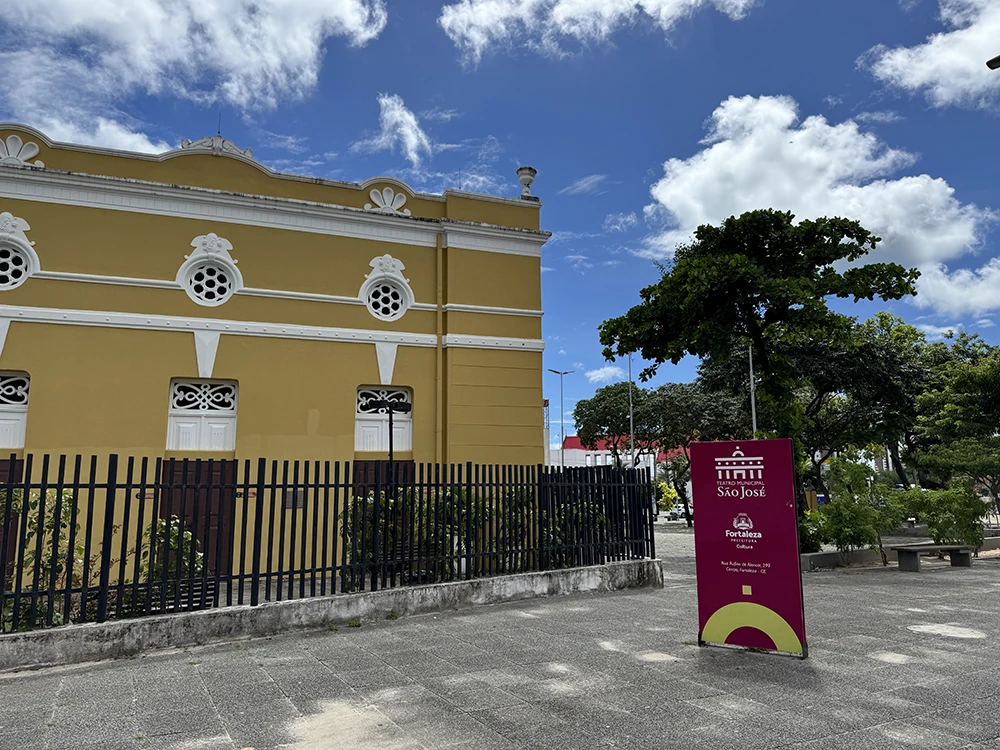 Fachada lateral do Teatro Municipal São José em Fortaleza, um prédio histórico amarelo com detalhes brancos, visto a partir de uma praça arborizada com céu ensolarado ao fundo.