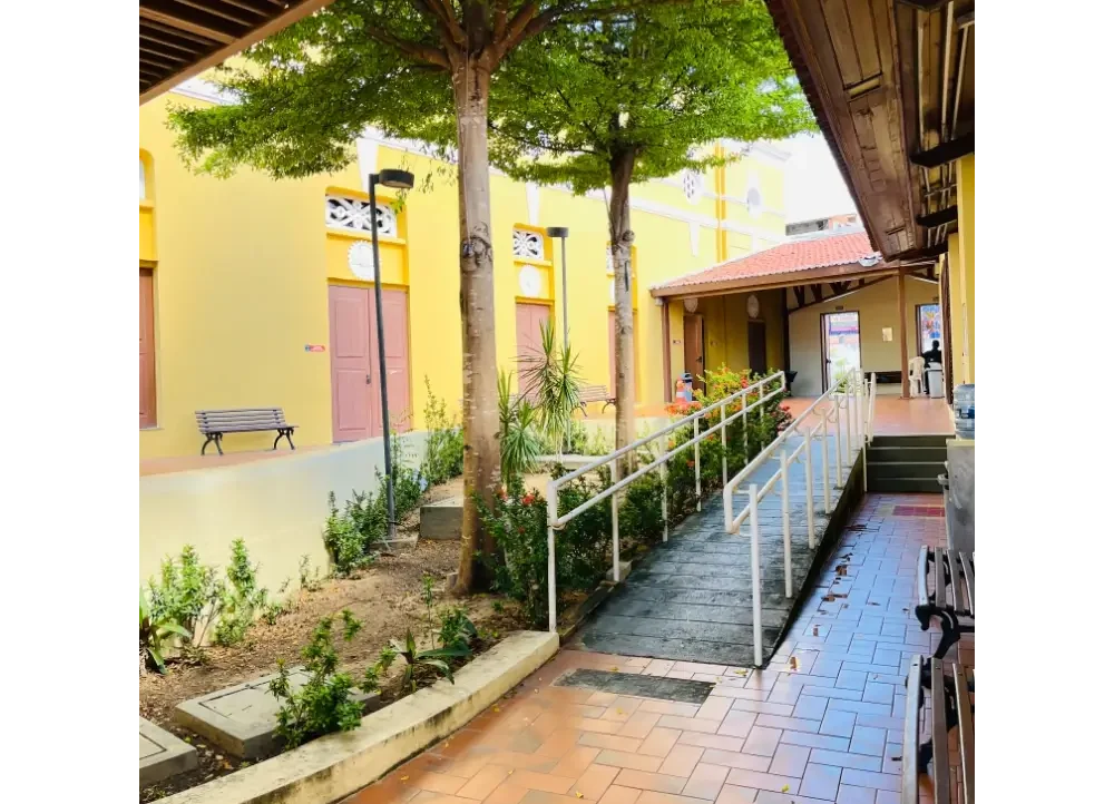 Área interna do Teatro São José em Fortaleza, mostrando uma rampa de acessibilidade com corrimão branco que leva ao pátio interno cercado por árvores e prédios amarelos